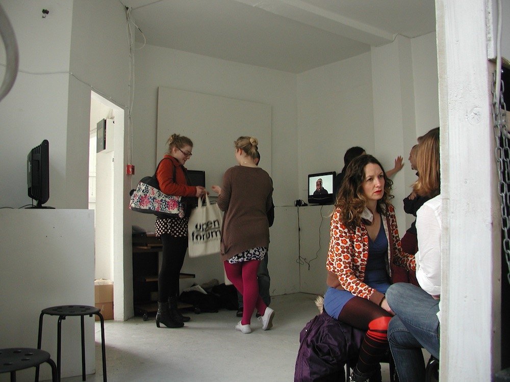 white project space with people talking and monitors screening videos