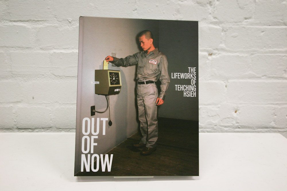 book cover of 'Out of Now: The Lifeworks of Teching Hsieh, Ed. Adrian Heathfield. On the cover, the artist Tehching Hseih, an American-Taiwanese person, stands next to a time clock in grey top and trousers. He is punching in his time card.