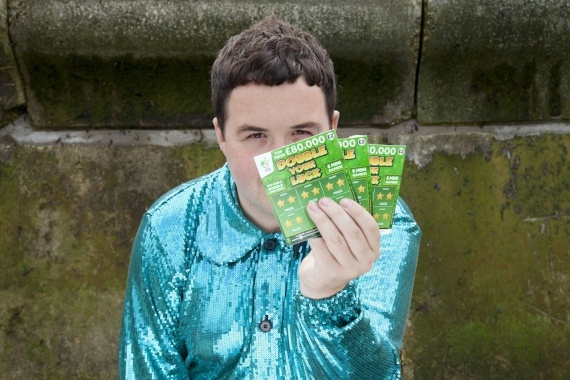 Scottee in a sequined turqouise shirt holding up three green cards in front of their face