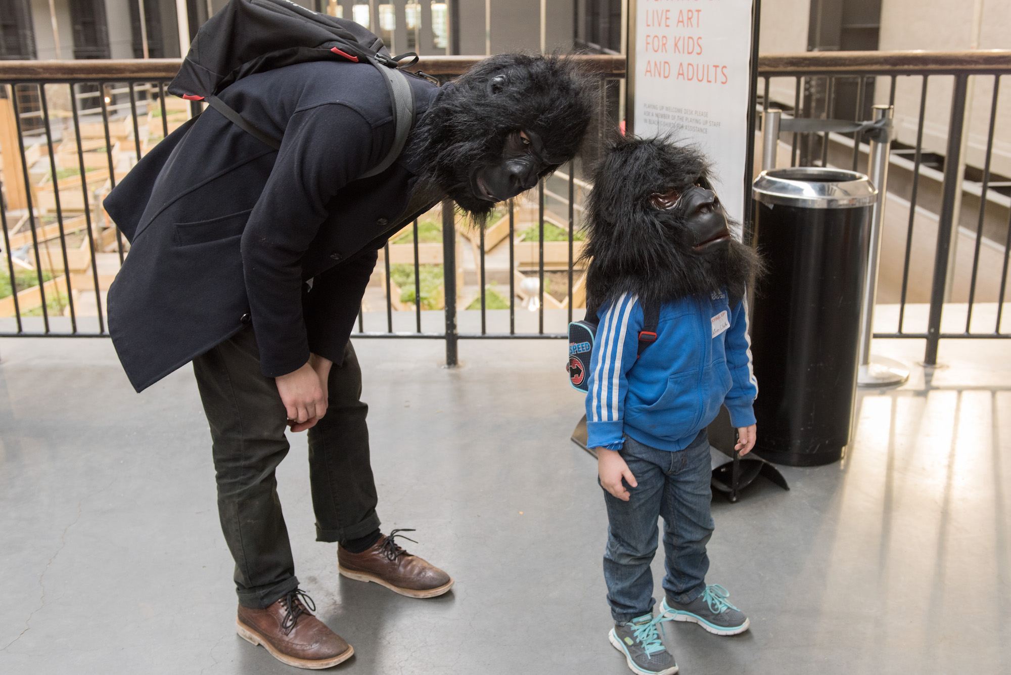 An adult dressed in a black coat wearing a gorilla mask leaning of a child in a blue sports jacket wearing a gorilla mask.