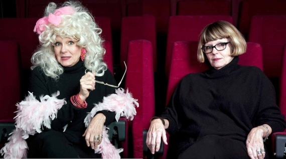 Artist is sitting on red seats. On the left the artist is dressed in their persona, wearing a pink feather boa, long earrings, bangles and a blonde wig decorated with a pink bow. On the right the artist is sitting as themselves dressed in all black garments.