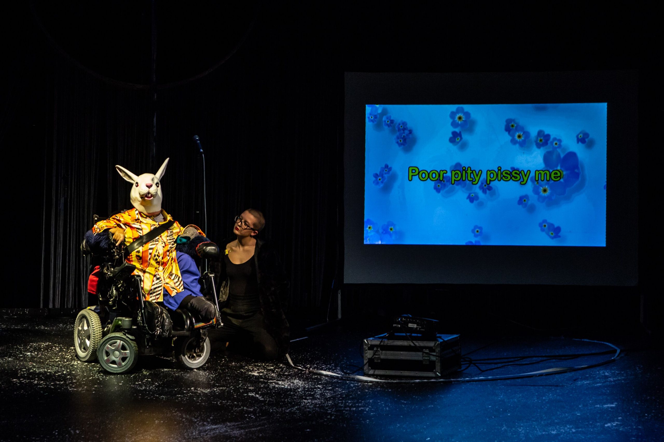 The artist is in a wheelchair and wearing a rabbit mask. To her side is her assistant. There is a screen to the right with the words 'poor pitty pissy me' projected.
