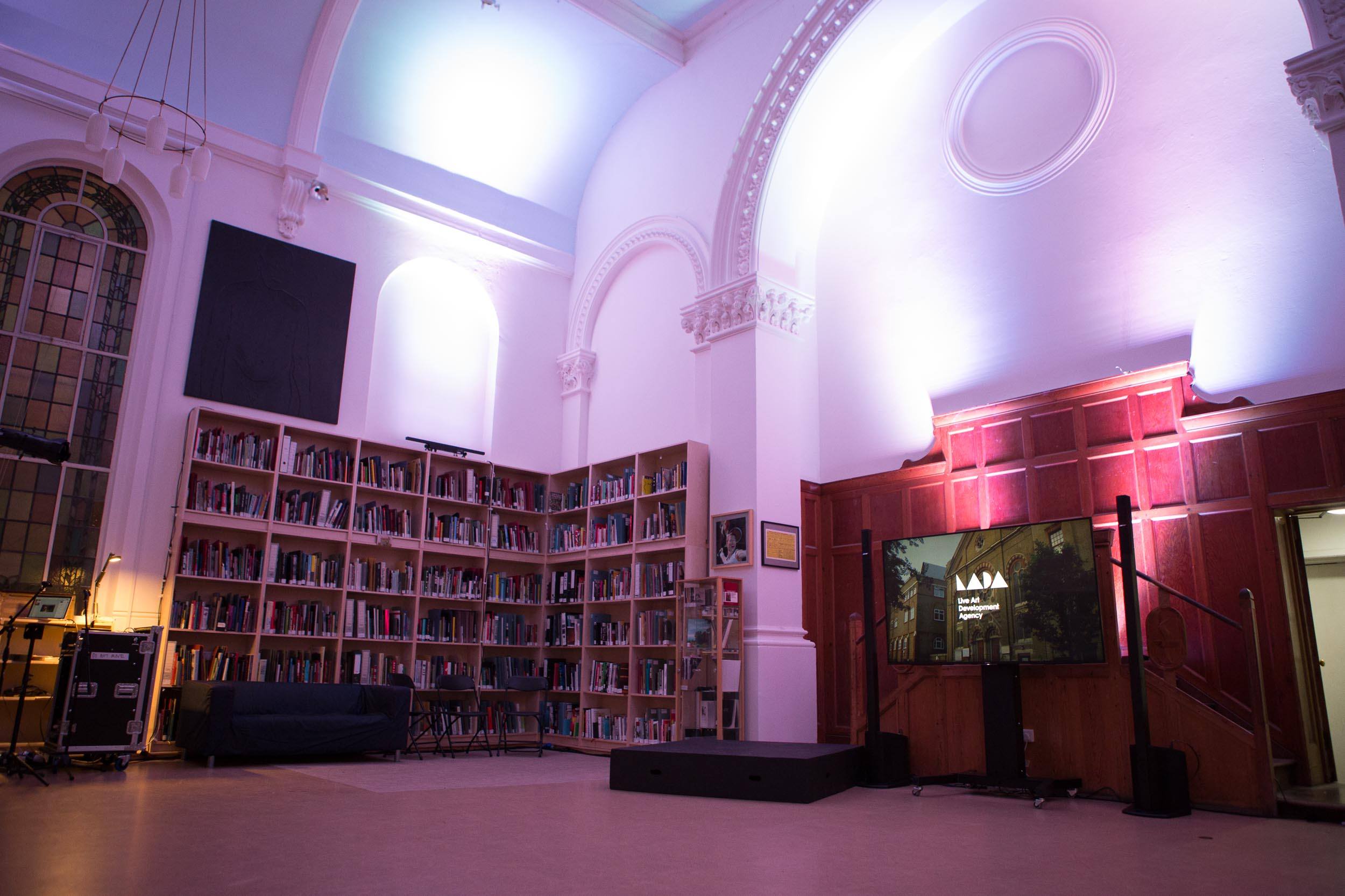 A low angle photograph of LADA's study room. The room has high ceilings and white walls. There are tall bookcases lining the walls and a large monitor displaying LADA's logo. The room is bathed in a pinkish light