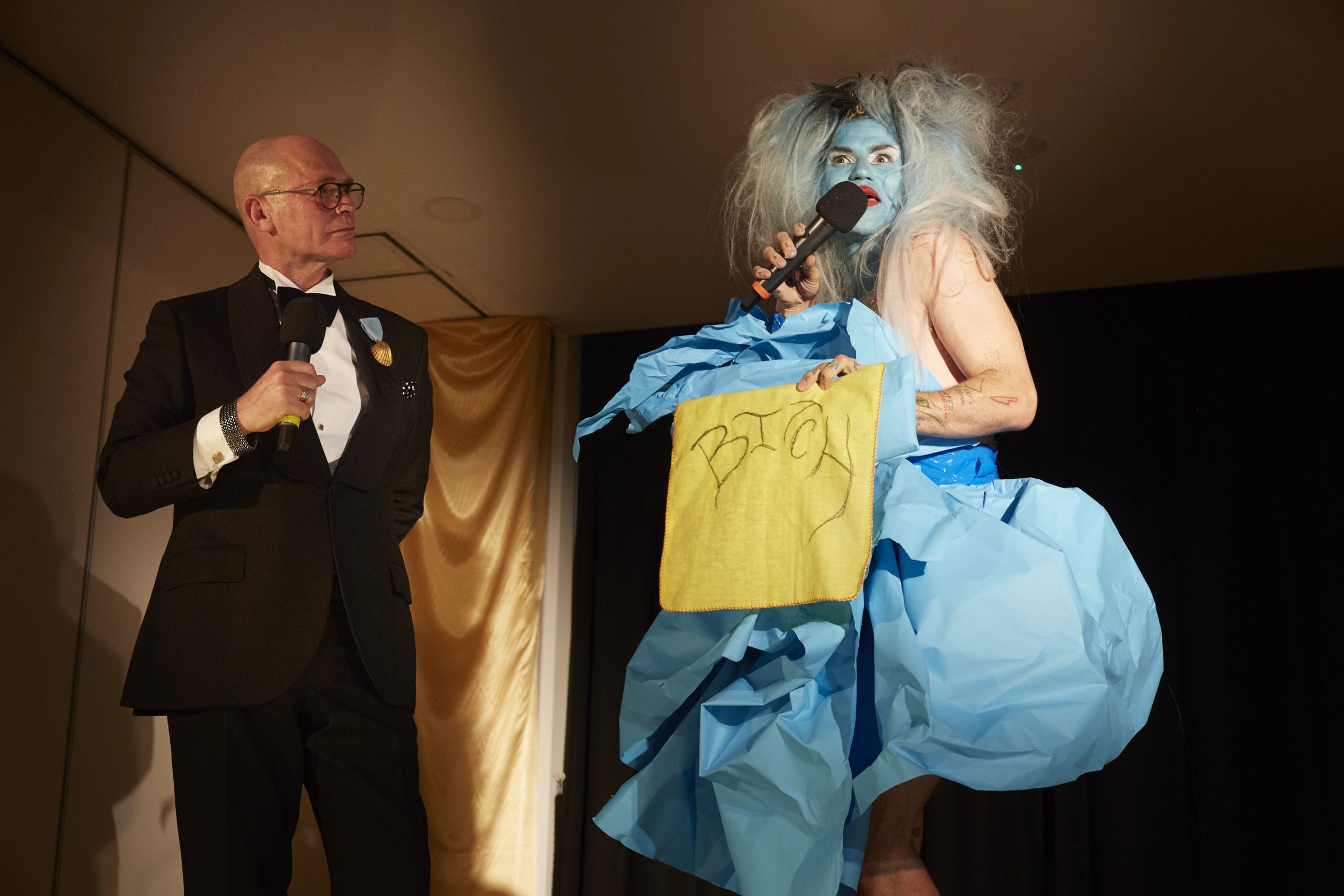 Drag performer with a blue face, wig and dress holds a yellow cloth with the word 'bitch' embroidered in it. An auctioneer in a tuxedo watches.