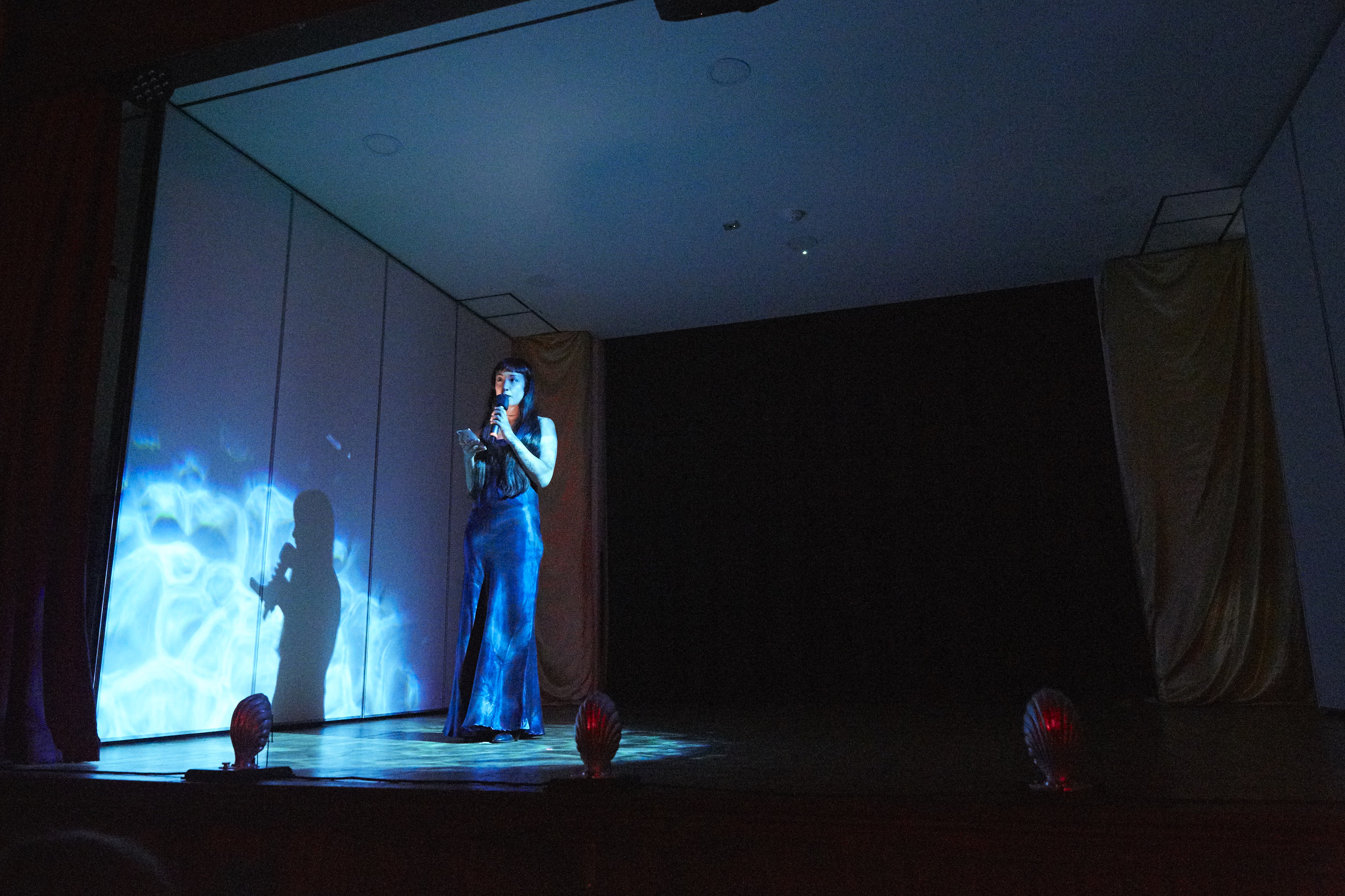 Artist reads a text into a microphone, lit by a rippling blue light with an effect similar to water.