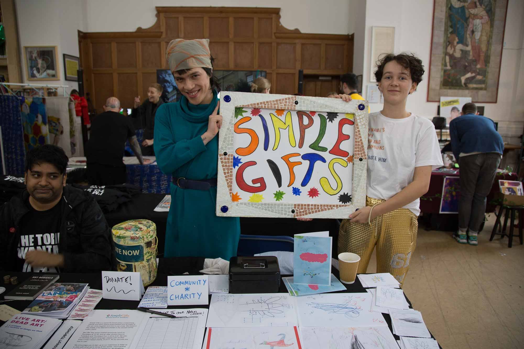 The simple gifts team hold a sign reading 'Simple Gifts' behind their stall.
