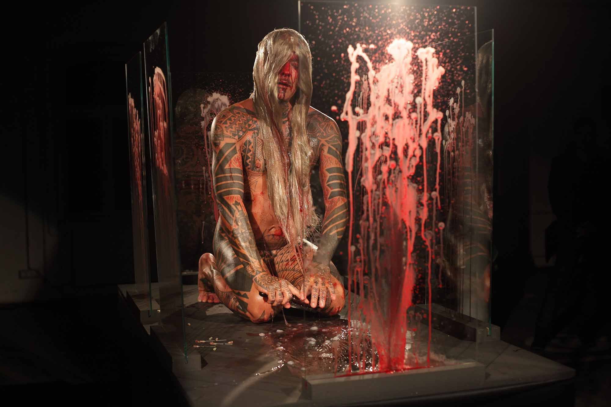 The artist is nude except for a blonde wig. He is surrounded by glass panes covered in his own blood.