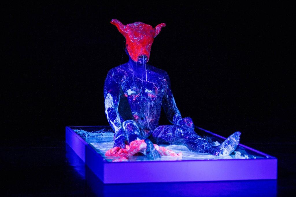 The artist is nude and lit by lights showing UV paint on his body. He wears a stylised minotaur mask.