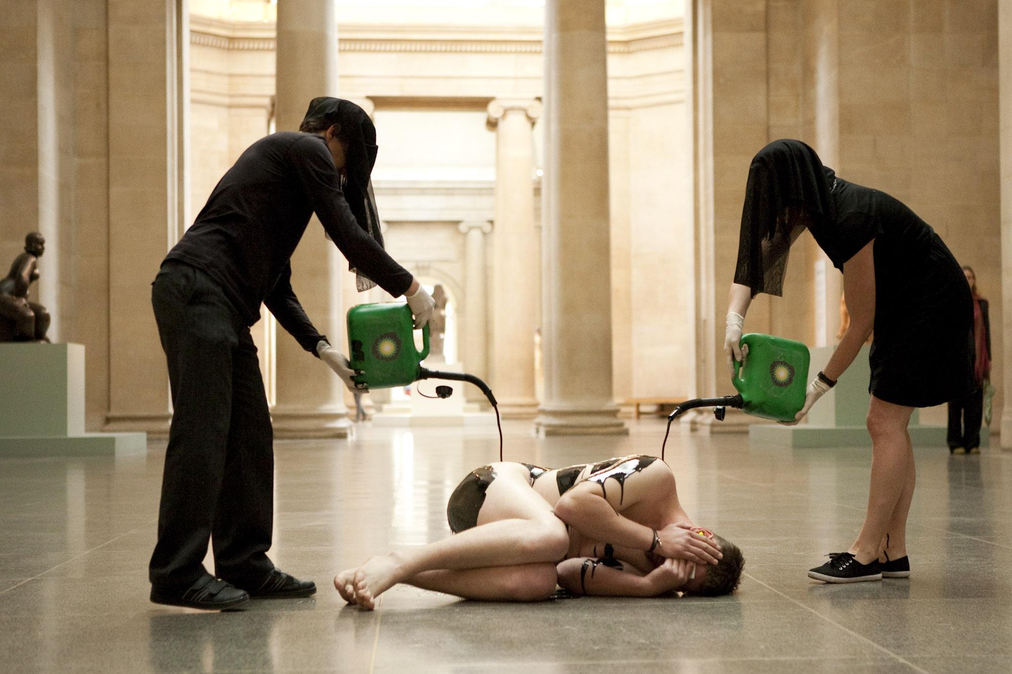 Two Liberate Tate activists pour a liquid resembling oil over a nude body in a gallery.