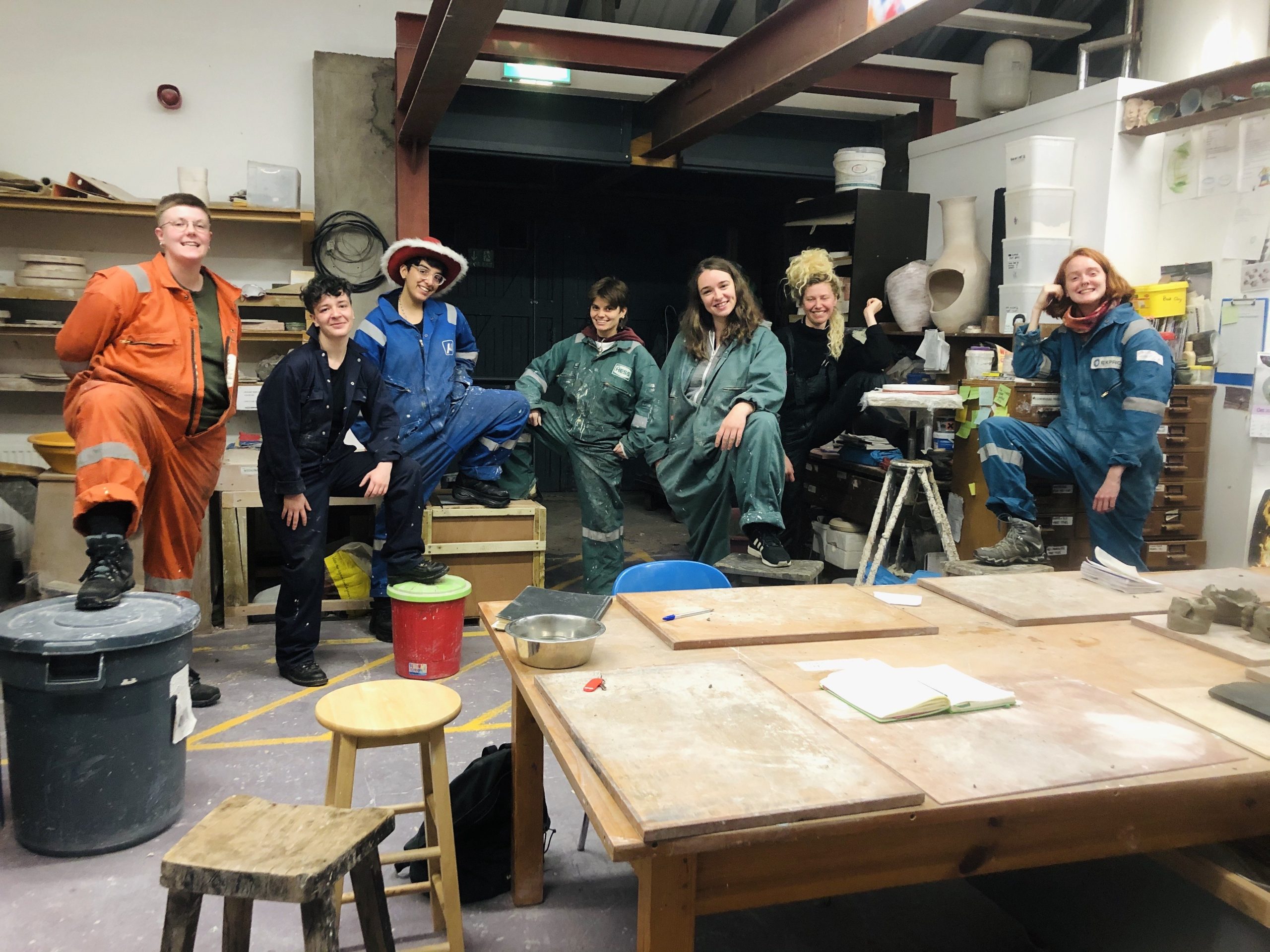 8 trans-masc artist pose in a clay/pottery studio in overalls, one person is wearing a red and white fluffy cowboy hat