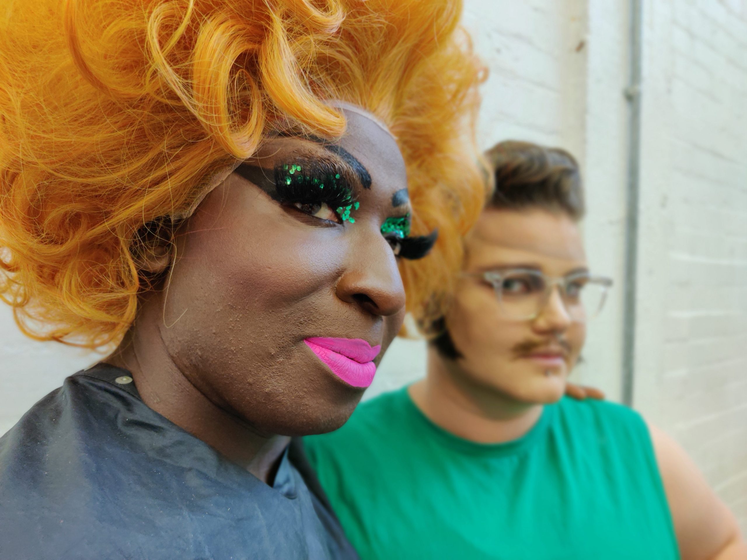 a close up image of a person in a large orange wig abd green glittery make up, with their arm around someone who is wearing a green t-shirt