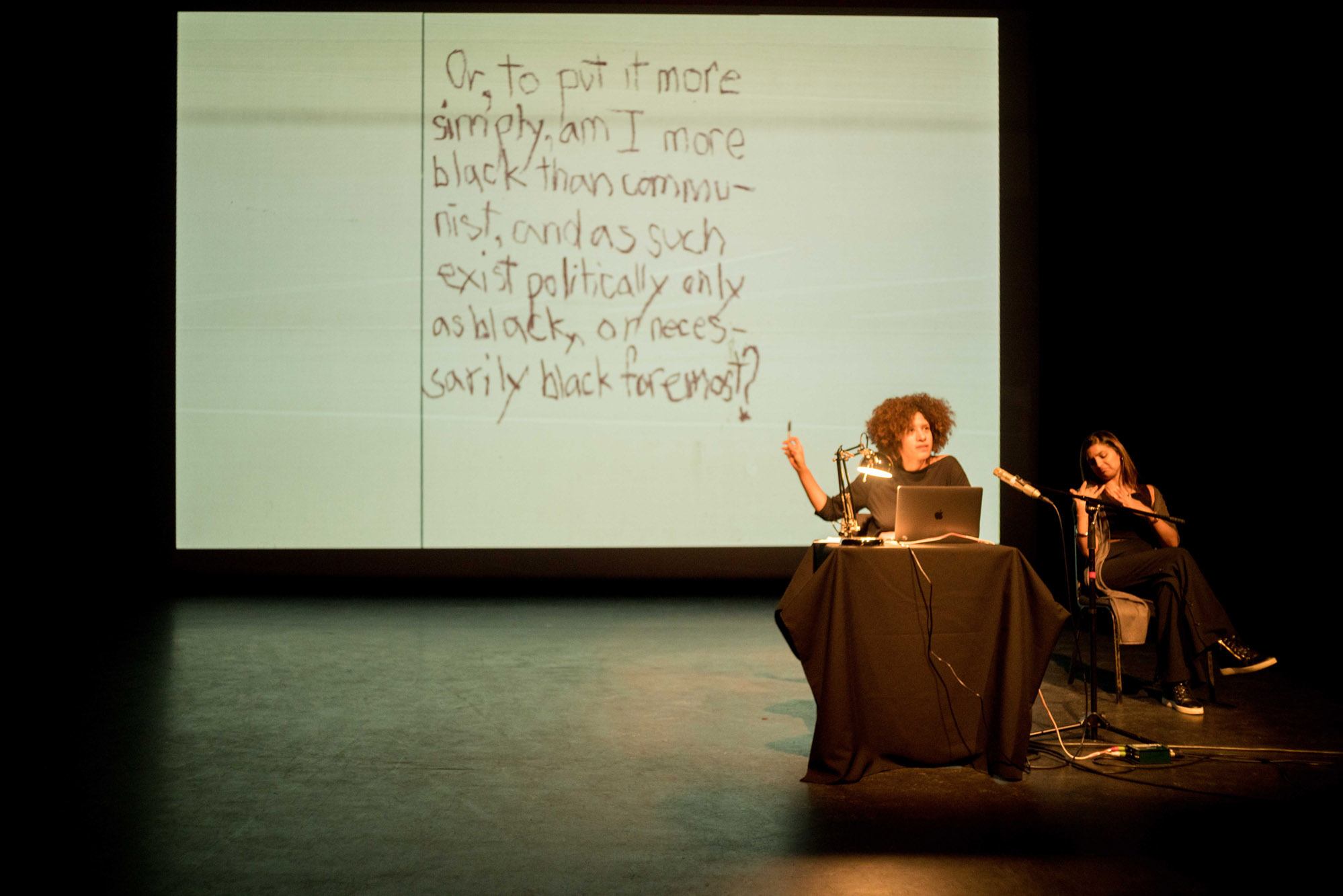 Performance: Season Butler (artist) sits at a table with a laptop and points to the projection screen behind her.