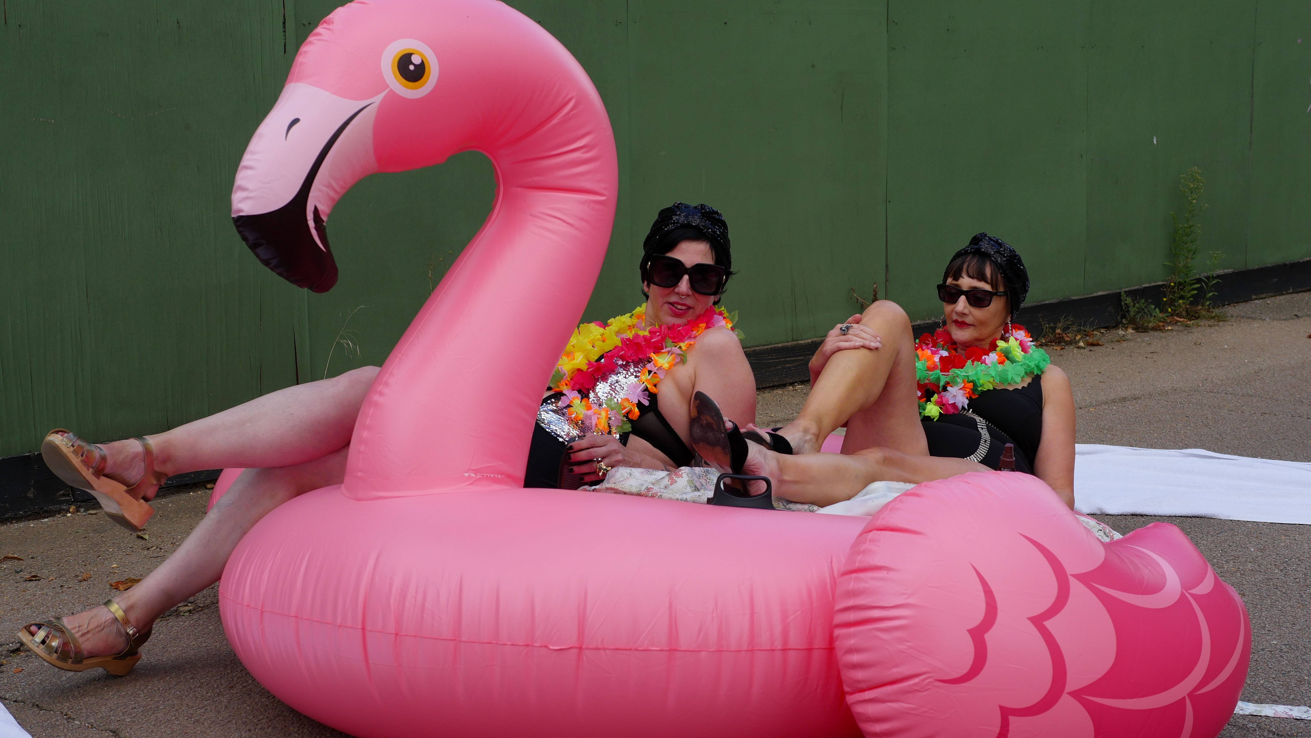 two women in lingerie recline leisurely in a blow up pini flamingo on a pavement