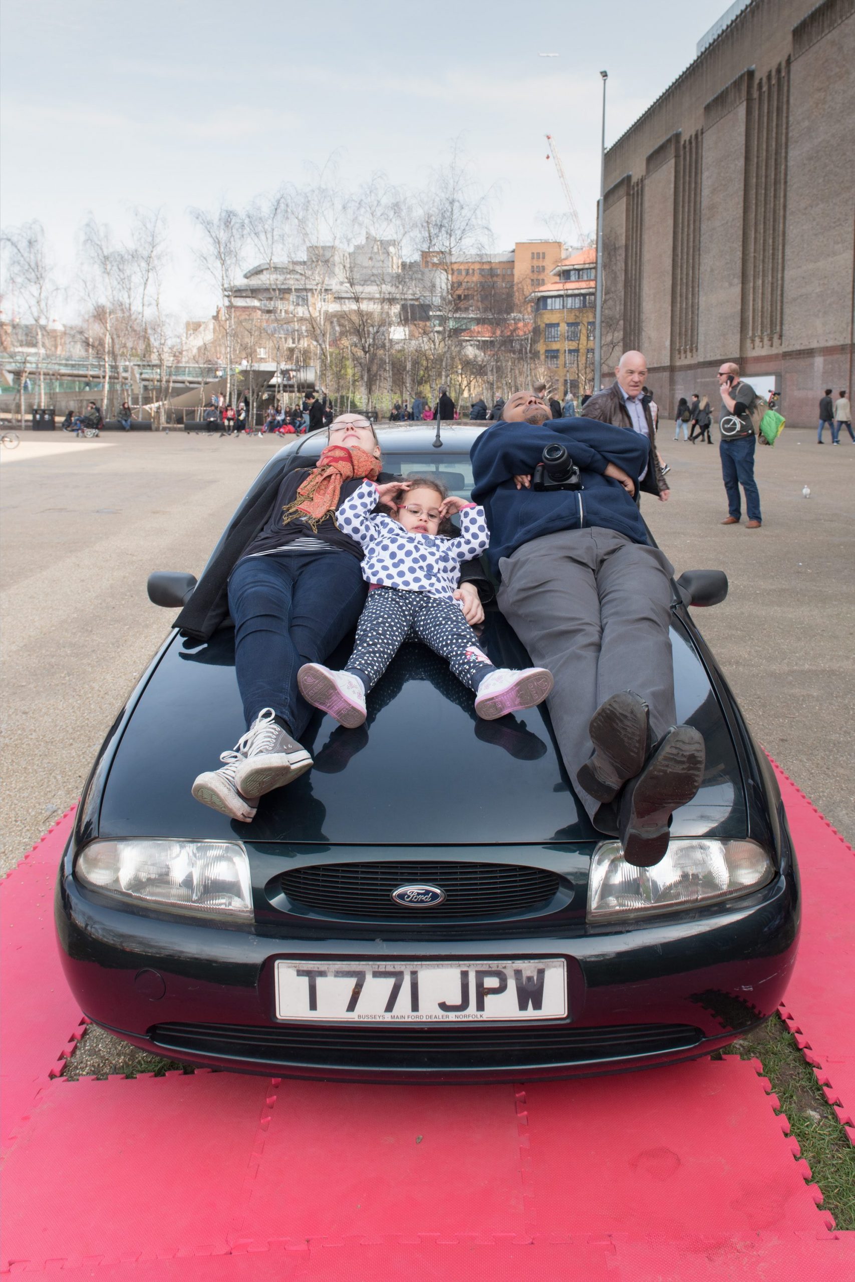 a child and two adults lie on top of a car outside the Tate Modern.