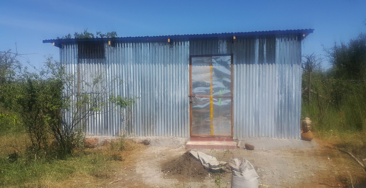 at the centre of the picture is a small house built with blue mabati sheets for walls, with a wooden-frame door. In the background, clear blue skyes and various vegetation and wild bushes surround the house. A dirt path leading to the front of the house.