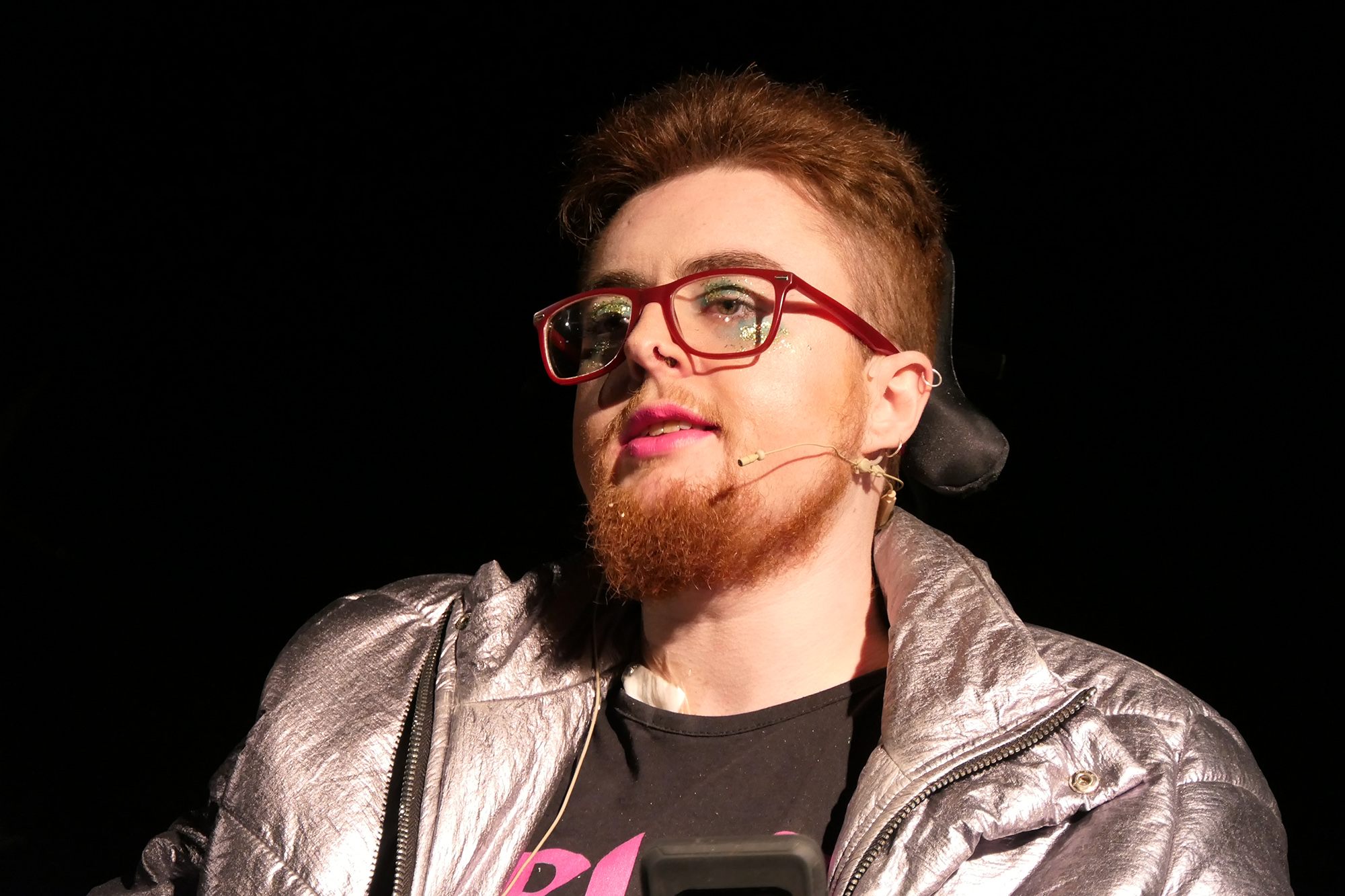A close up of the artist, a young queer person in a wheelchair, wearing eye shadow and a silver jacket.
