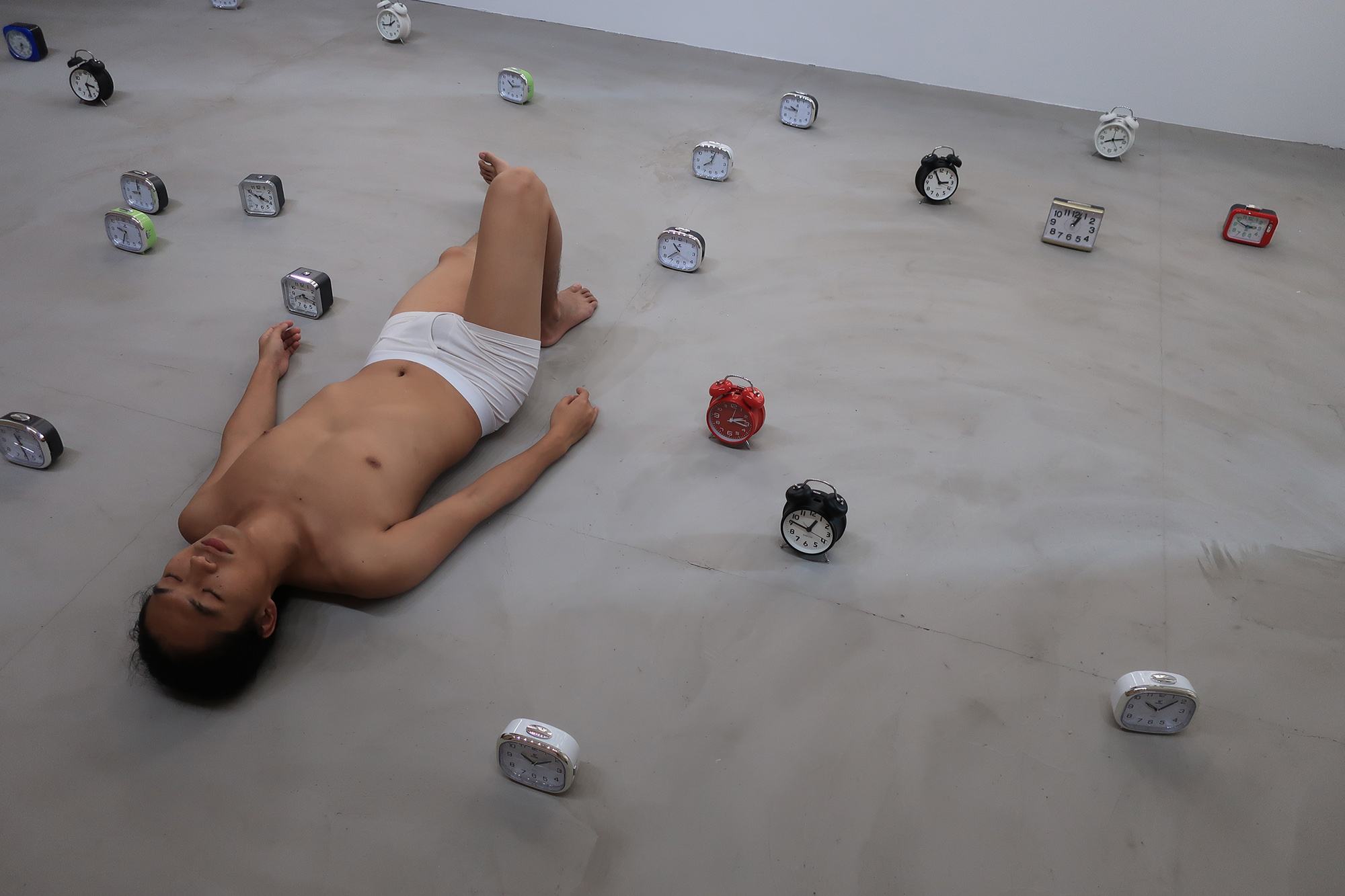 The artist, a young Taiwanese man, reclines in his underwear in a gallery of alarm clocks.
