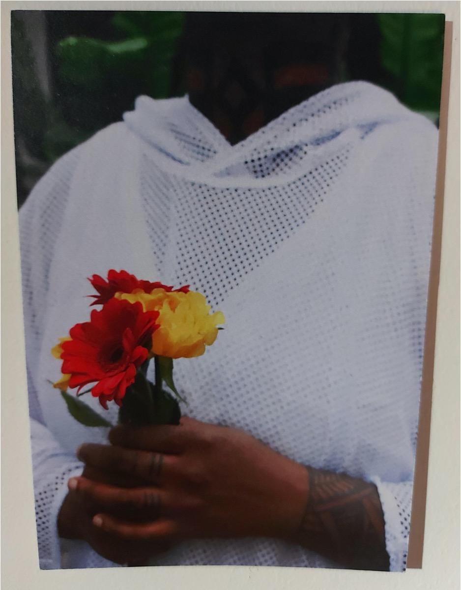 A close-up of a person in a white mesh hoodie holding flowers. They are a black person with a tattoo showing on their wrist and fingers.