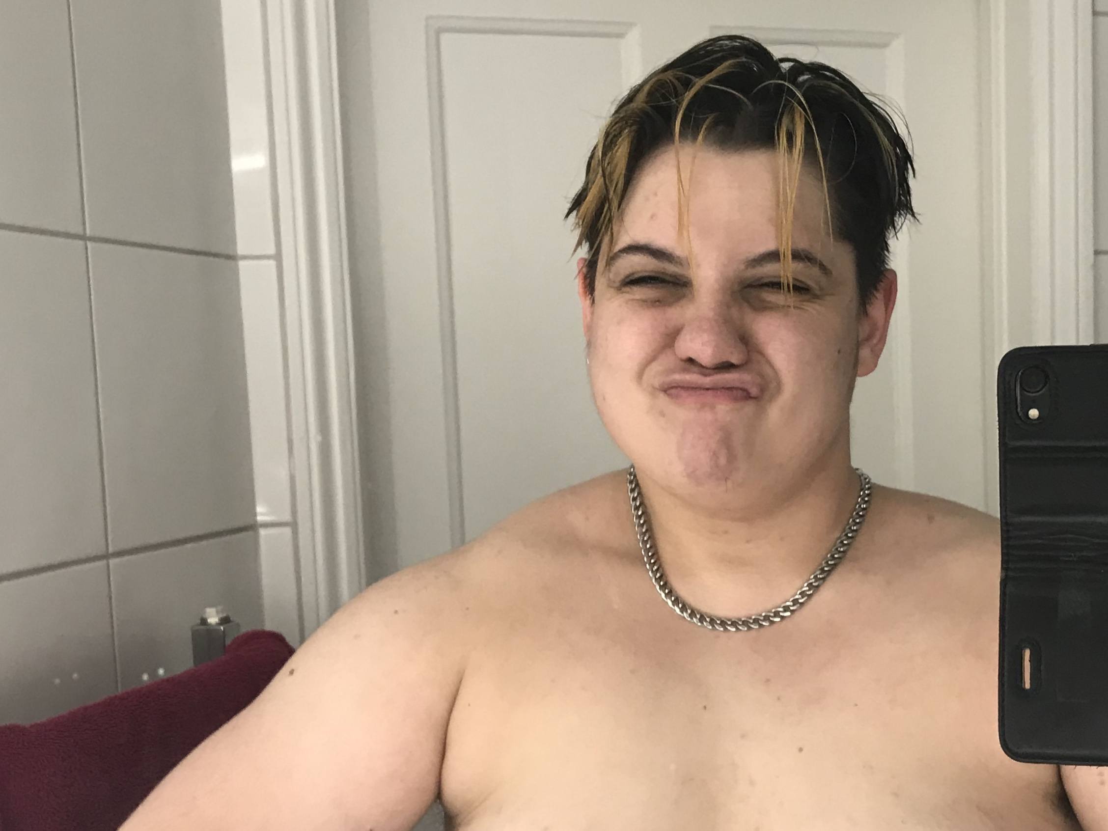 A selfie of Rabi, standing in a white-tiled bathroom topless making muscles in the mirror. He is a young, mixed-race, fair-skinned, transmasc guy with short floppy black hair with gay blonde streaks. He has a playful pout and wet hair, fresh from a bath. He is wearing a heavy silver chain.