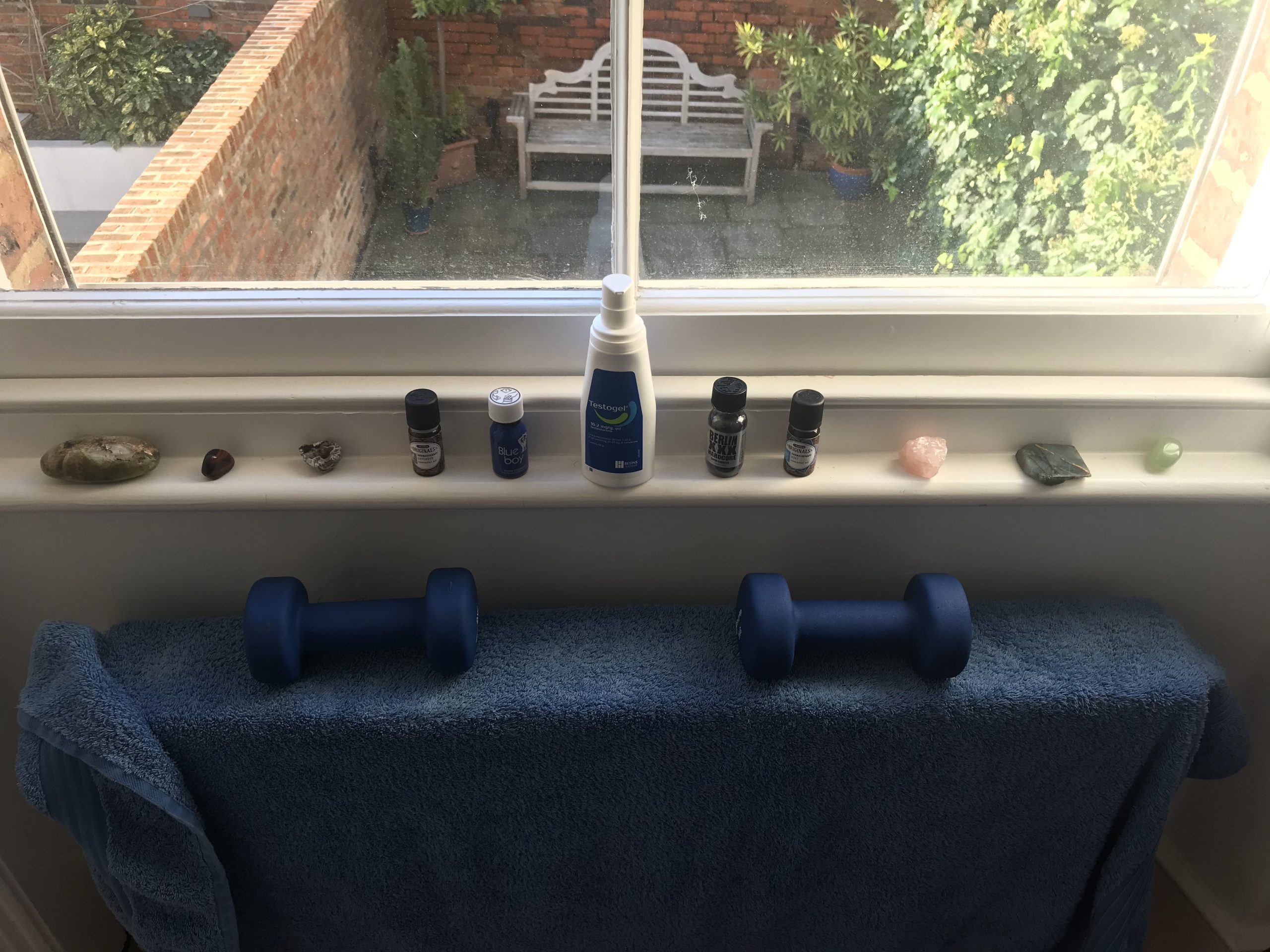 An alter: a bottle of testogel on a thin window ledge with either side of it various small bottles and crystals. Below is a blue towel on a radiator, weighted down by two blue free weights. The image is sunny, and out the window at the top you can see a terrace garden below with a wooden bench and some greenery.