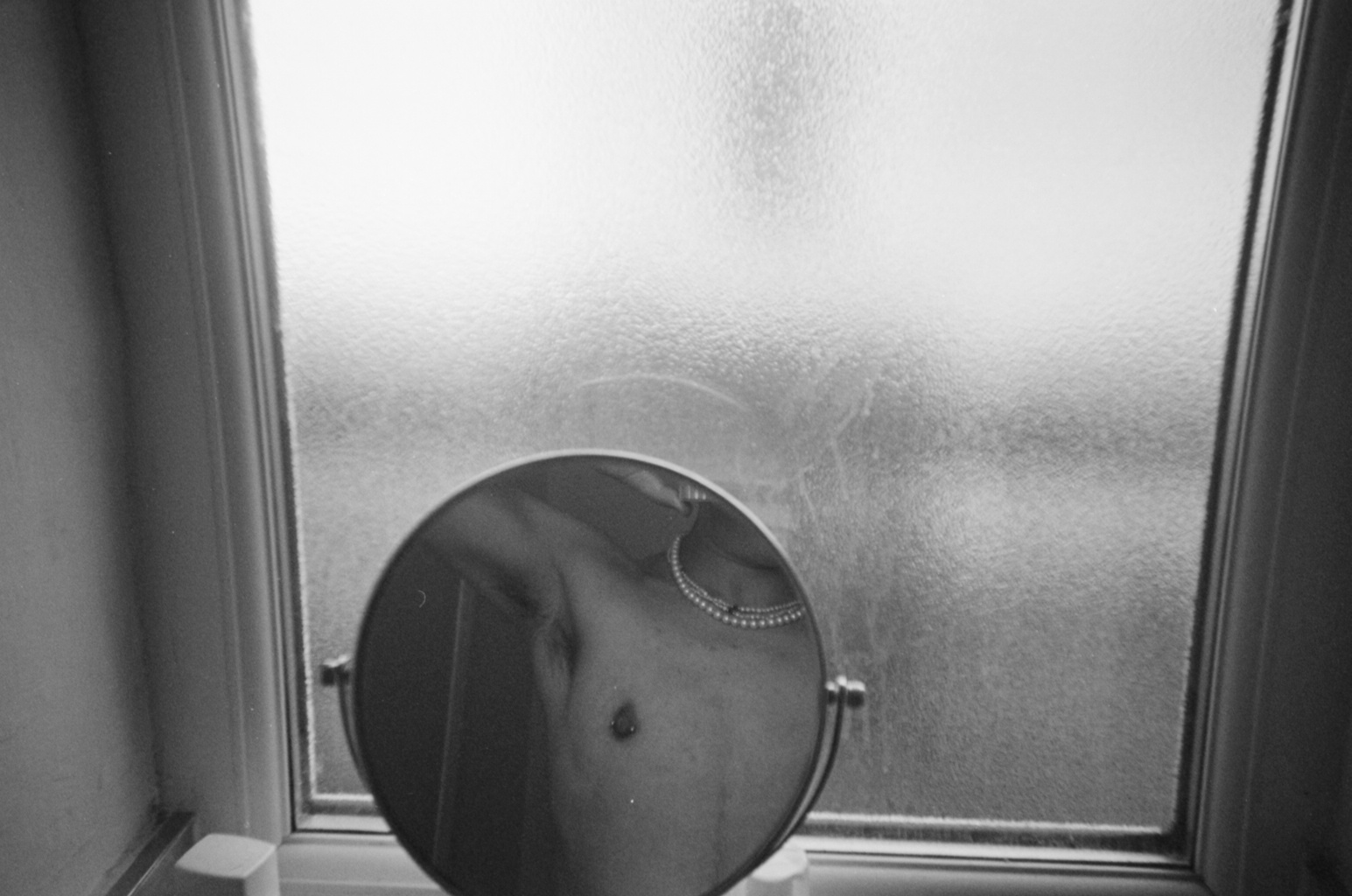A black and white image of a small circular mirror infront of a window. The window has frosted glass, obscuring the view. In the small mirror a torso is reflected. The person wears two pearl necklaces and lifts their arms behind their head