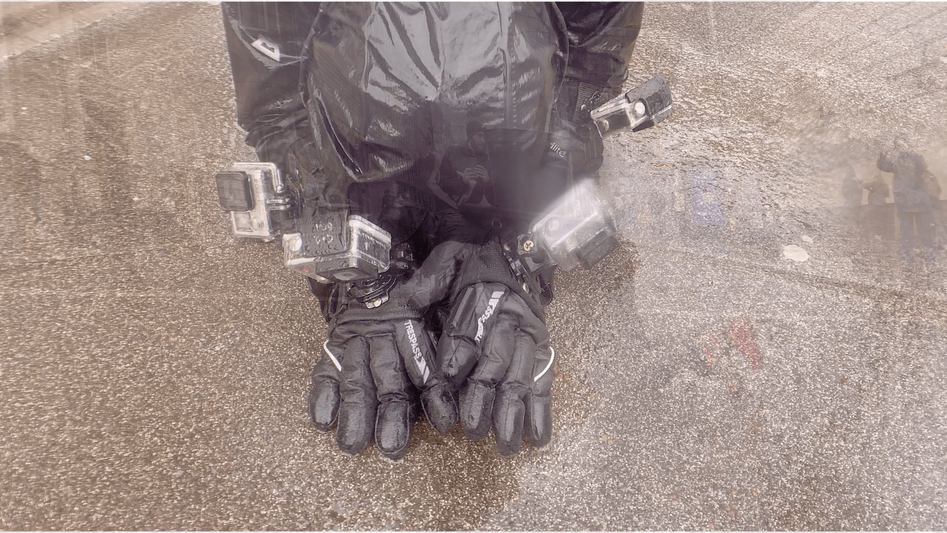 A woman has let her head drop onto her forearms, her black gloved hands are stretched out on a tarmac surface. The gloves have the brand logo ‘Trespass' written on them. Four GoPro cameras are attached to her hands and wrists. The image has been overlaid by another image, so on her wet black hood you can see an image of a man holding a phone camera and the plant pot and the tree trunk, there are also some very faint and so difficult to identify colours and shapes from the overlaid image in the wet pavement.