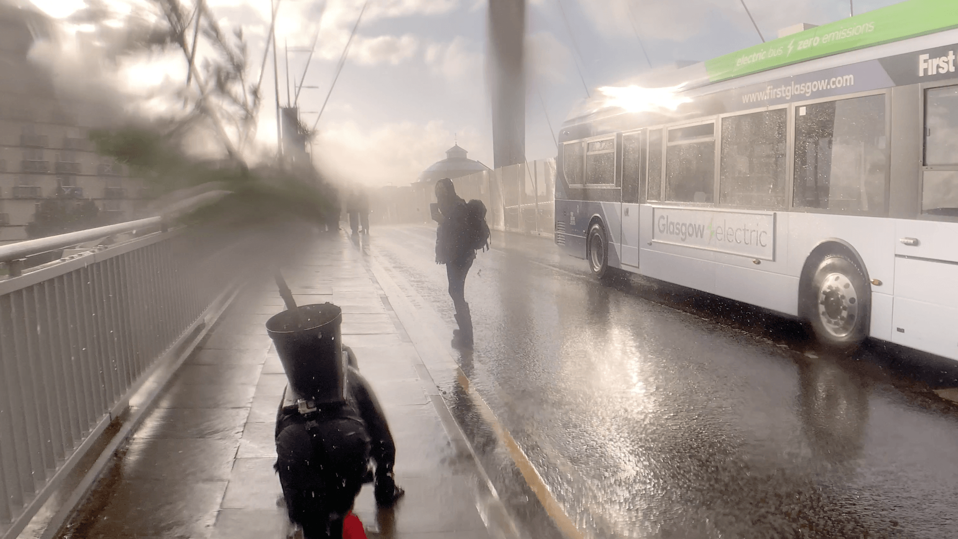 A woman is crawling away from the camera on her hands and knees. She is wearing black waterproofs, knee pads, black gloves and red trainers. She is carrying a large black plant pot on her back with a Scots Pine tree in the pot swinging towards the right of the image blown by the strong wind. She is crawling on a wet paved pedestrianised pavement on a bridge. A bus is passing her on her right on the other side of the road, ‘Glasgow Electric’ is written on the side along with other advertisements and a website link. It’s a very active image with strong contrasting light, rain has landed on the camera lens creating blurs and distortions. There is a strong light flash on the bus and drops of rain spray on the lens. A woman is standing to the right of Miranda, on the road filming with her phone camera.