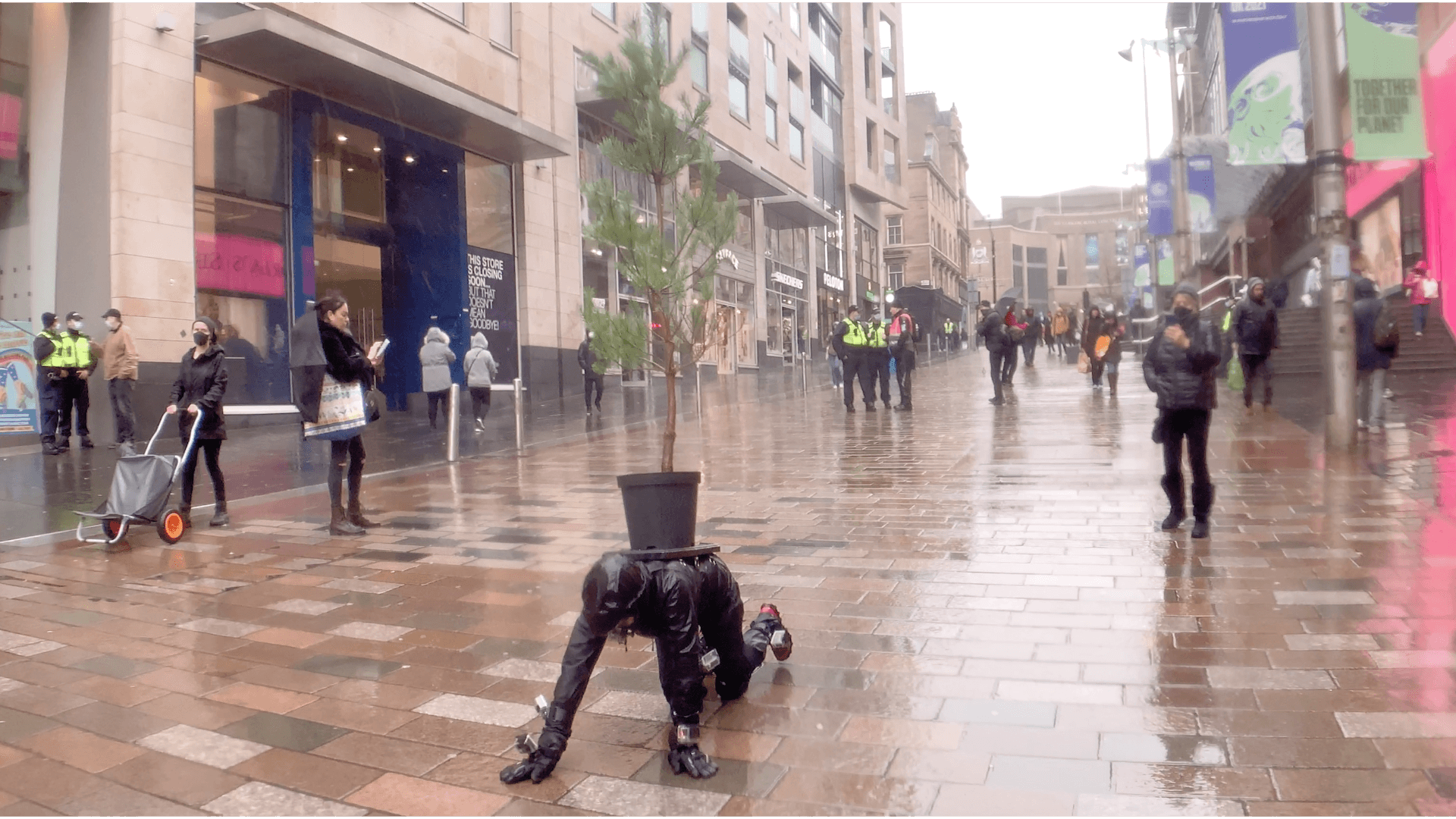 A woman is crawling towards the camera on her hands and knees, on a wet paved pedestrianised shopping street. She is wearing black waterproofs, knee pads and black gloves. Her hood is up so we cannot see her face. She has GoPro cameras attached to her arms, hands, wrists, legs and there is one in her mouth. She is carrying a large black plant pot on her back, there is a Scots Pine tree in the pot. It is pouring with rain, many people are wearing face masks. Groups of security guards wear high vis yellow jackets.