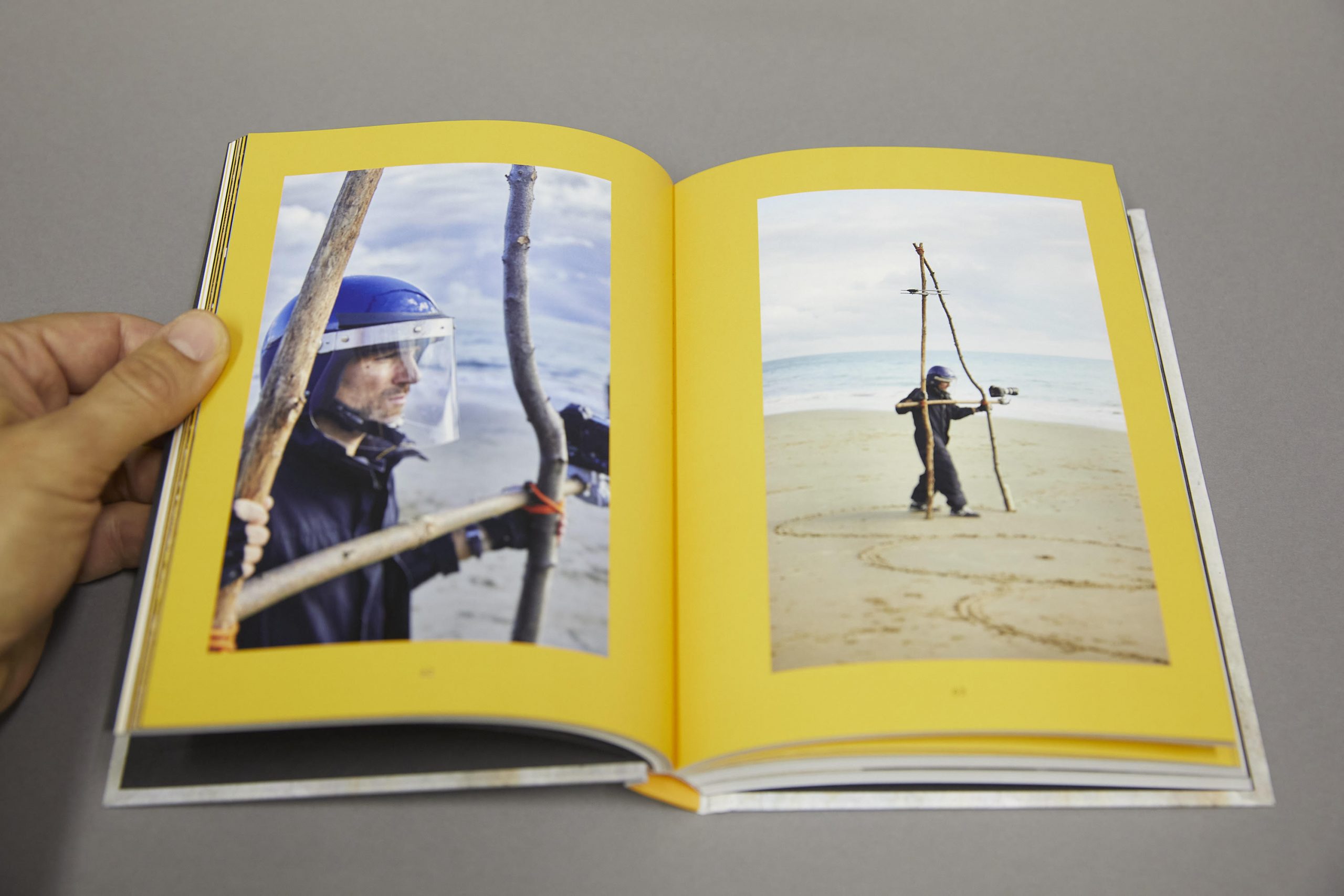 Photo of the book Unframing Photography on a grey surface with a hand flicking through it. The two pages we see feature two large photos surrounded by yellow frames. The photos depict a white man, wearing workwear and a blue helmet and using a wooden pair of compasses to draw circles on a sandy beach. The pair of compasses has a camera attached to it.