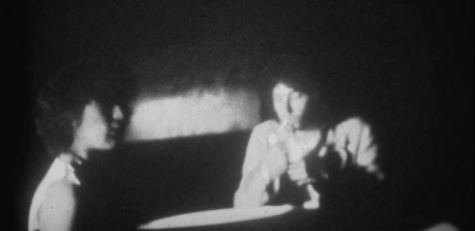 A blurry black and white photograph of two people. The person on the left is in profile, with half their face in shadow. The person on the right is facing towards us, looking down at the cigarette they are lighting