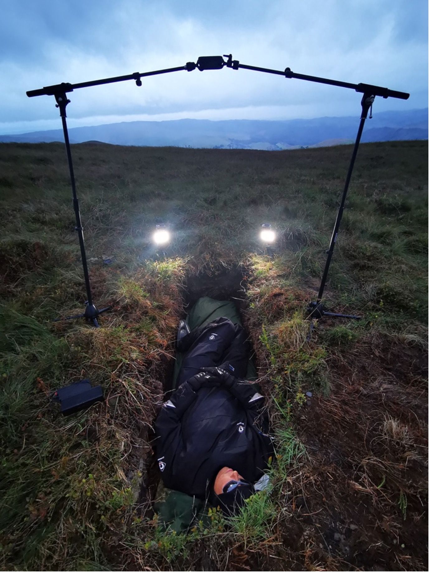 Miranda Whall laying in a 2 foot self-dug ditch at night time, with two lights above her illuminating the hole