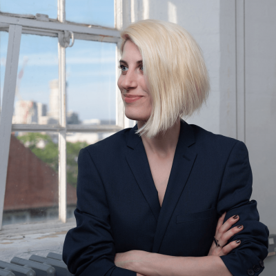 Natalia Damigou-Papoti, is a white female with shoulder length blonde hair. Natalia is stood wearing a black top, she has her arms crossed and is looking thoughtfully smiling out of a window on a sunny day