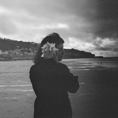 A black and white shot of Rosaleigh Harvey-Otway, who is a white woman with short black hair and wearing a black jumper. She is stood on an empty UK beach, listening to a large shell to her ear with her eyes closed.