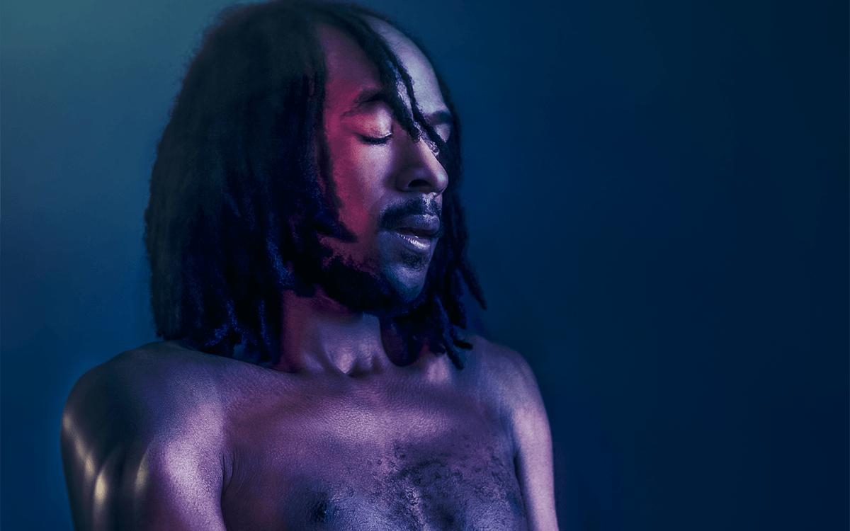 Jamal Gerald, who is a Black man with shoulder length dreadlocks, is in a blue-lit room with his eyes closed whilst dancing. A purple light is lighting up his naked body.