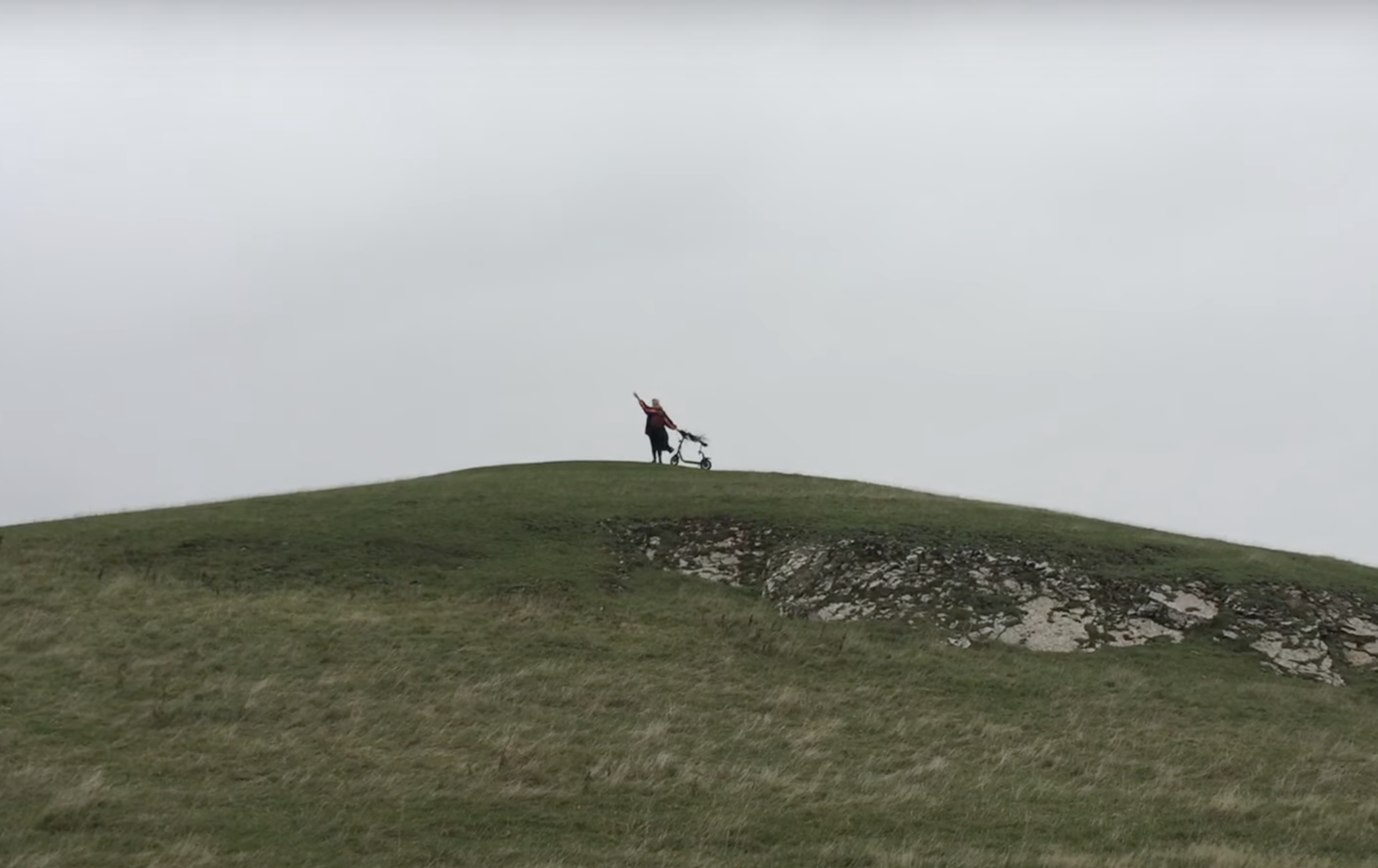 A person is on the top of a hill, waving at the camera and holding a mobility scooter with their other hand.