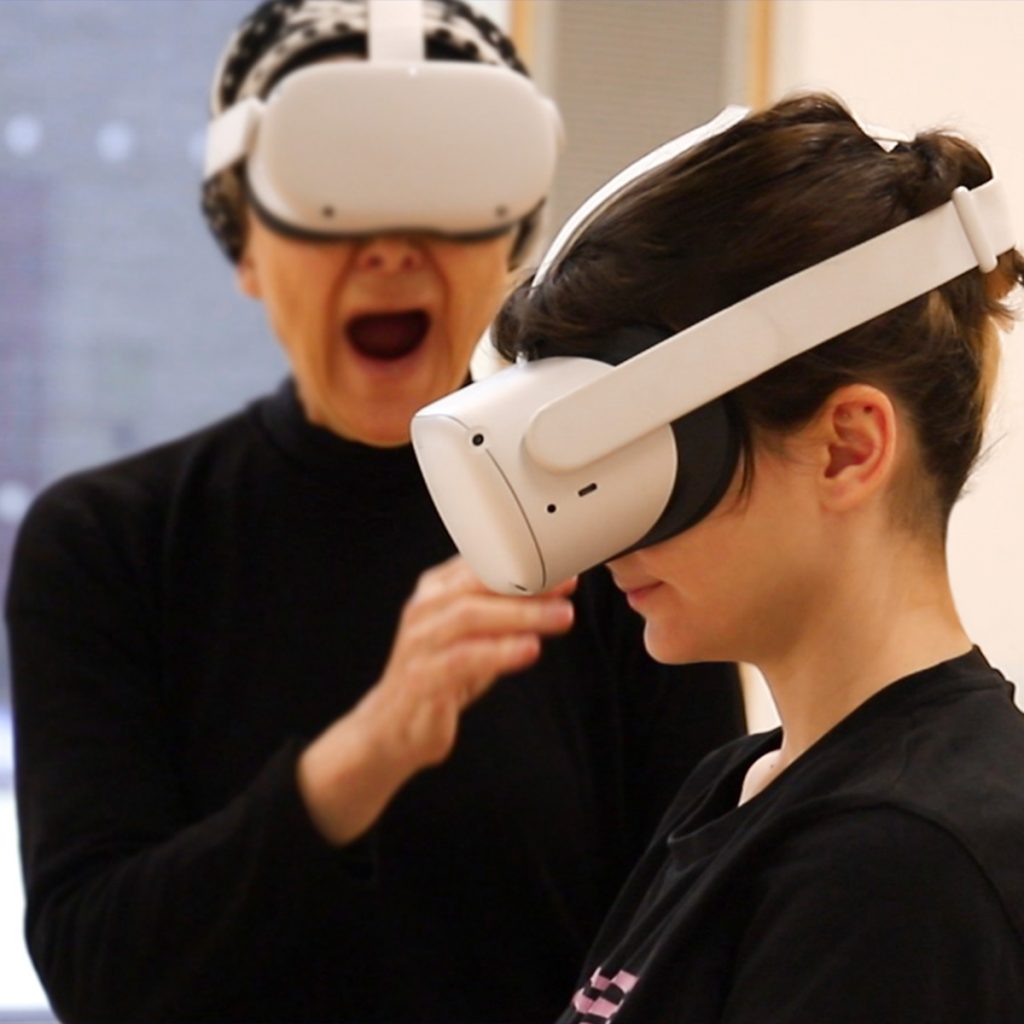 Two pale skinned people wearing VR headsets and black tops. One is facing the camera, with their mouth open and then other is side on with their head slightly down, listening.
