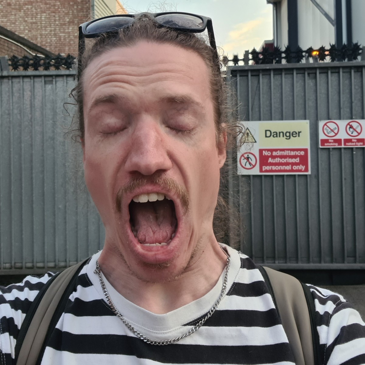 A white person wearing a black and white striped shirt takes a selfie with their mouth wide open and eyes closed, standing in front of a gate marked with warning signs.