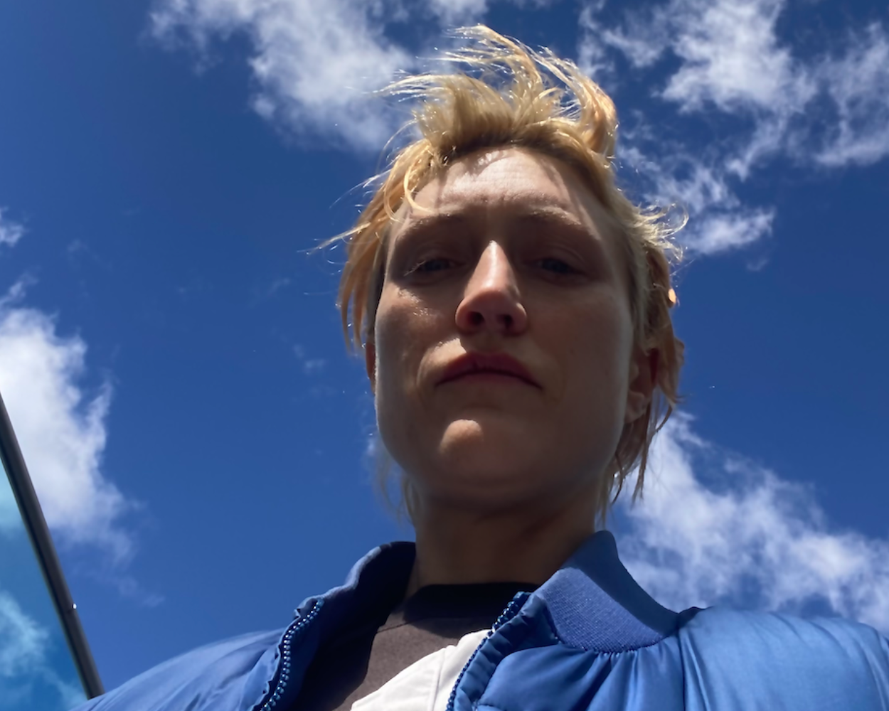 A white person with short blonde hair looks down at the camera under a bright blue sky with scattered clouds, wearing a blue puffer jacket and layered clothing. The image has a dramatic upward perspective with natural lighting.