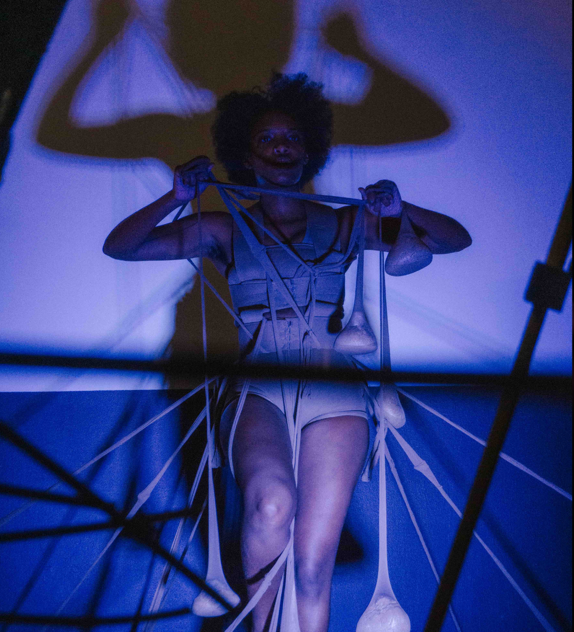 A black person in a harness-like outfit is entangled in long cords with weights. Their shadow is projected behind them. The photo is dramatically lit in blue.