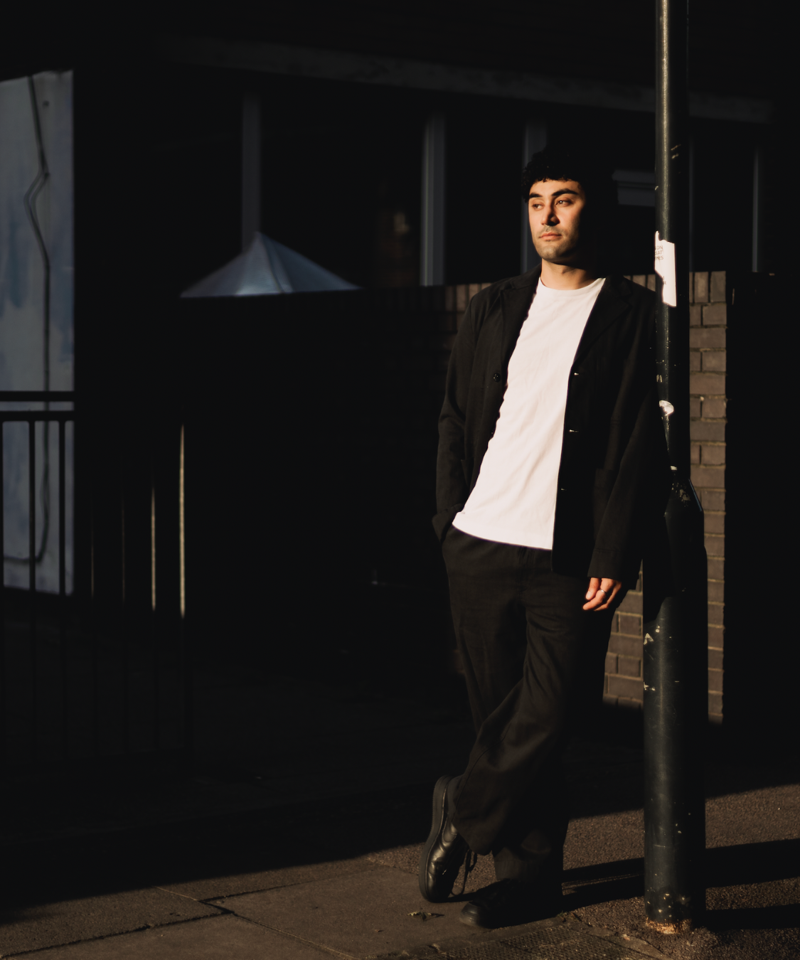A person in a black outfit and white shirt leans against a lamppost on a dimly lit street, partially illuminated by a beam of light. The background is mostly in shadow, creating a dramatic urban atmosphere.