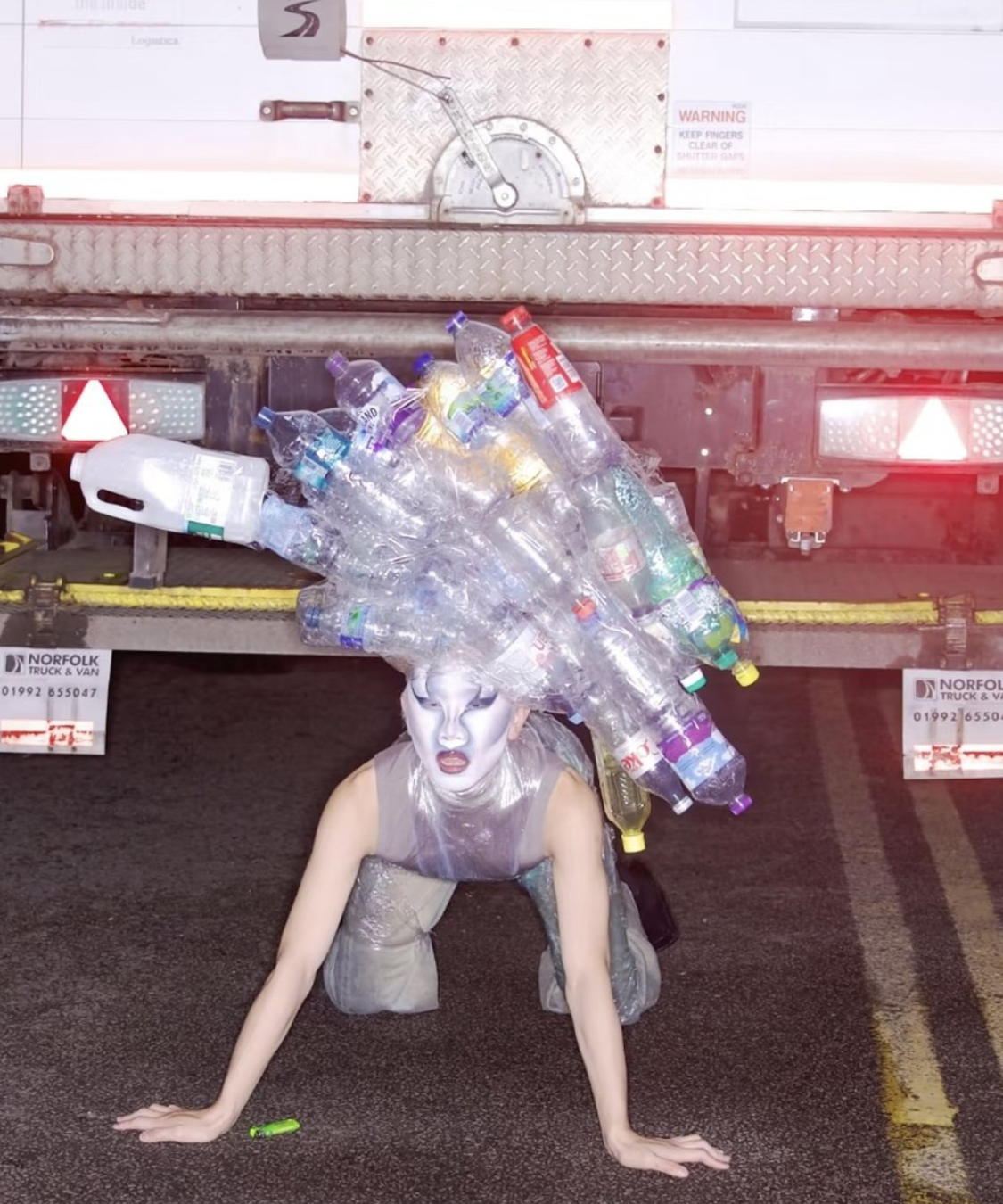 A person crawling on the ground beneath a truck's rear. Their costume features a headpiece made of empty plastic bottles and plastic waste and their body is wrapped in transparent material resembling plastic wrap. They’re wearing a full face of alien-like drag makeup.
