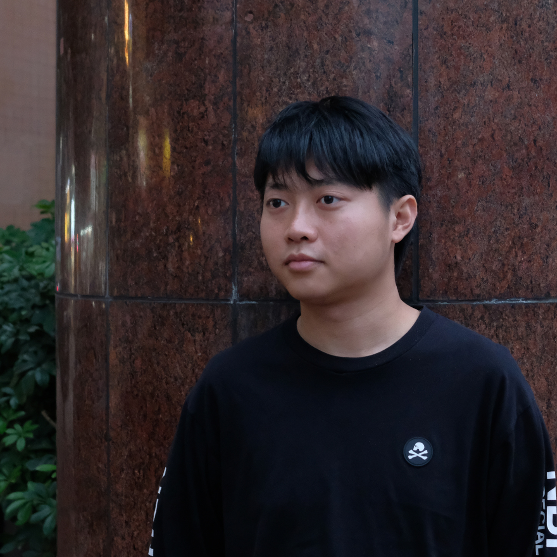 A person with short black hair stands in front of a polished brown stone wall, wearing a black shirt with a small skull emblem.