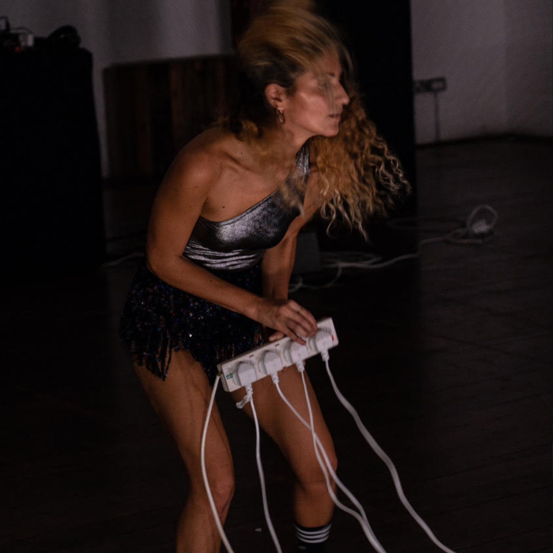 A person with olive skin and curly long hair that is dark at the roots and blond at the ends Is in motion holding a 4 way extension plug. We can see 4 plugs plugged in with cables that are out of shot. She is wearing a tight one shoulder silver leotard, a short sparkly fringed skirt, Adidas socks and trainers. Behind Claudia is a table with DJ equipment that is out of focus.