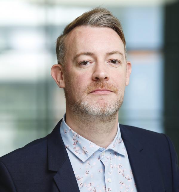Portrait of a blonde light-skinned white man. He's wearing a dark blazer and a pale blue floral shirt. He's staring at the camera and his background is faded.