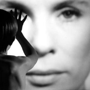 Black and white film still showing a person from behind, raising their hand toward a large, blurred face projected in front of them. The figure’s silhouette contrasts sharply with the close-up of the face.
