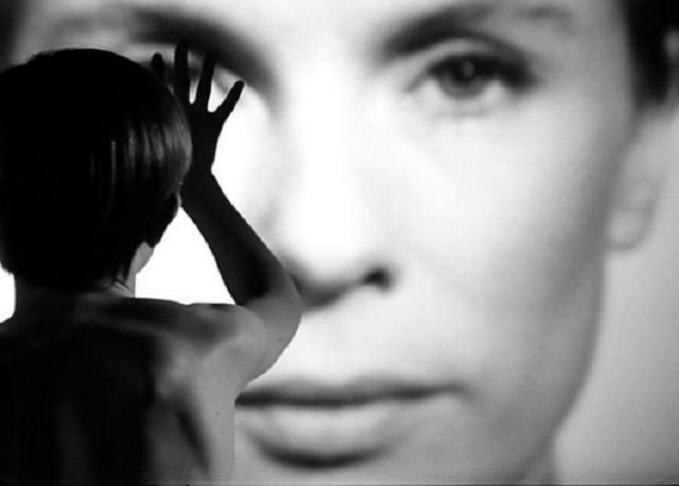 Still from Ingmar Bergman's Persona (1966). Used under CC Black and white film still showing a person from behind, raising their hand toward a large, blurred face projected in front of them. The figure’s silhouette contrasts sharply with the close-up of the face.