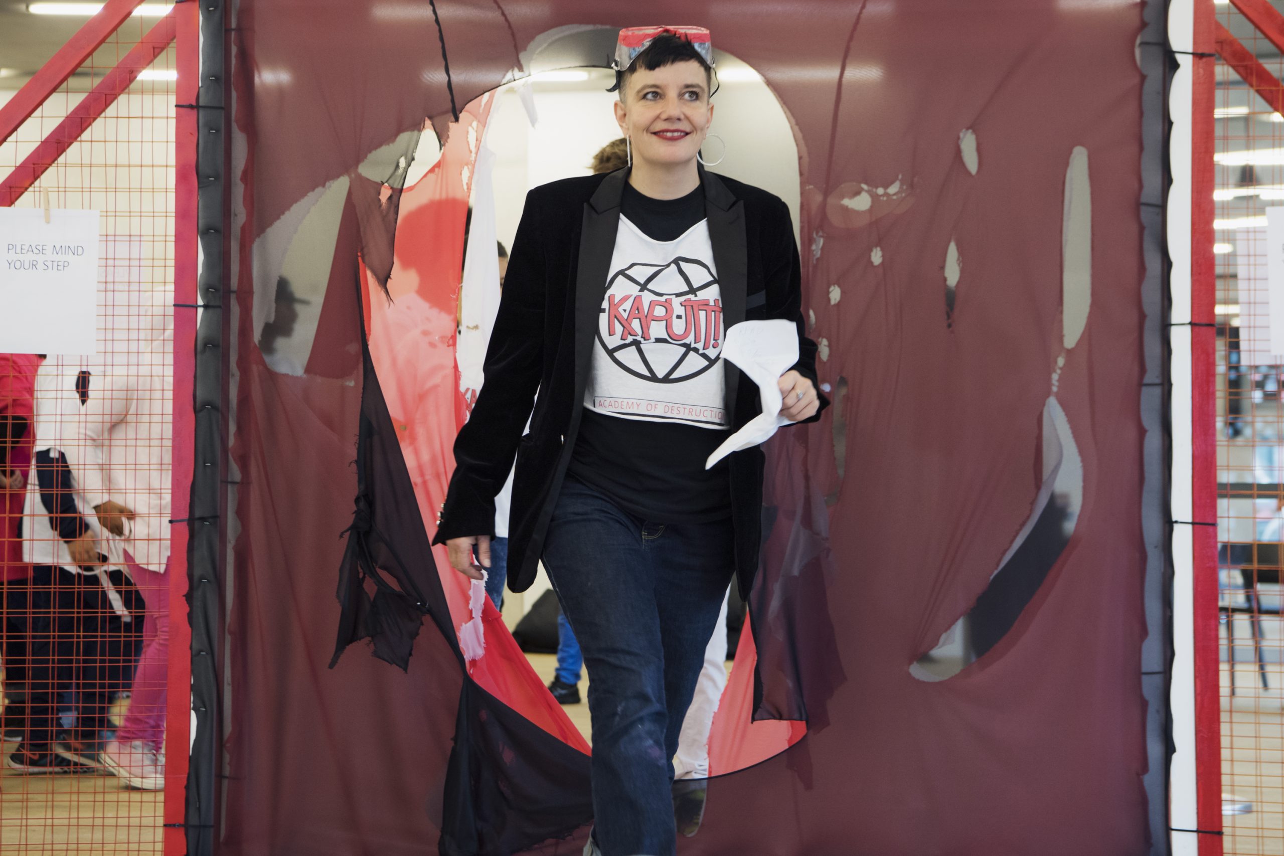 A white person wears a black suit jacket, jeans, a white t-shirt that reads “Kaputt - Academy of Destruction”, goggles on their head and large hoop earrings. They are walking towards the camera with ripped mesh behind them.