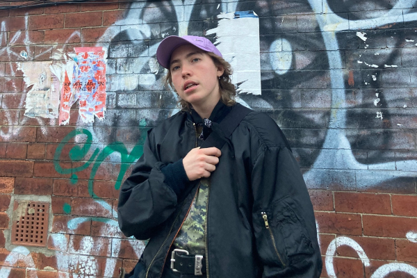 A photo of kane stonestreet in front of a wall with graffities. They're wearing an oversized black bomber jacket and a light purple baseball hat and look at the camera