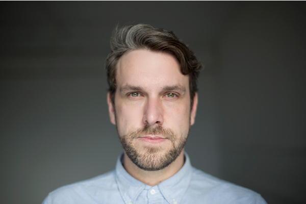 Professional headshot of a man with greying brown hair and beard wearing a light blue collared shirt against a gray background