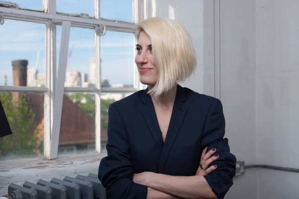 Natalia Damigou-Papoti, is a white female with shoulder length blonde hair. Natalia is stood wearing a black top, she has her arms crossed and is looking thoughtfully smiling out of a window on a sunny day